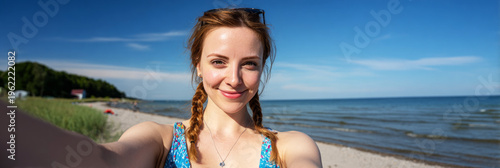 Girl with braided hair smiles while taking a selfie on a sunny beach. The sea is in the background along with a clear blue sky. People enjoy the beach on a bright day