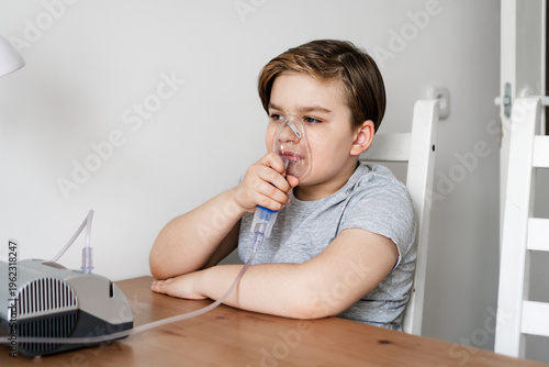 Young boy sitting at table using nebulizer for breathing treatment. Child receiving respiratory care and treatment at home. Concepts of childhood illness, respiratory treatment, home care.