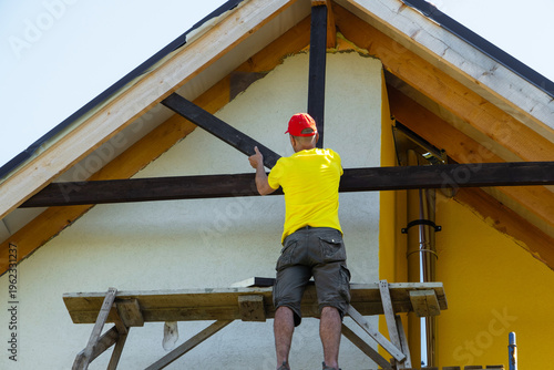 Photos Male carpenter attaches crossbeam to wooden house gable with screwdriver, white and yellow wall, focus on craftsmanship, height risk