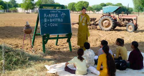 Indian young adult gen z female lady talk speak teach to villager child day under tree rural area. Happy girl boy group sitting on mat do study look board learn read english at open class field place
