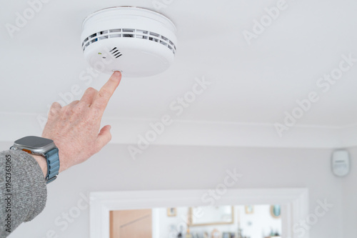 Handyman seen doing a weekly test of a combine smoke and carbon monoxide tester in a home hallway. The detector is wired into the house wiring a requires no battery.