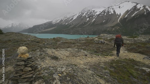 Hiker walking along a rocky path near a mountain lake under a cloudy sky in nature