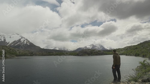 Hiker standing by a mountain lake looking at snow-capped peaks under a cloudy sky in nature