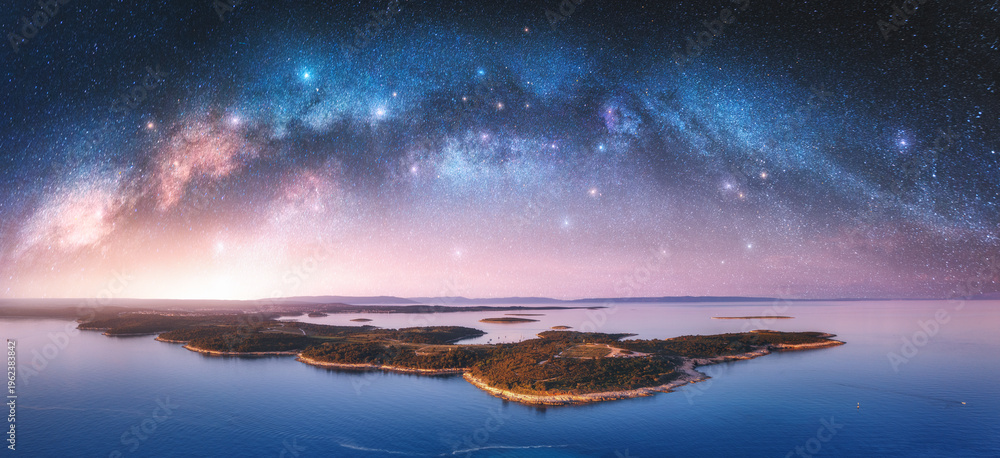 Fototapeta premium Milky Way arch and small islands in the sea at summer night. Kamenjak cape, Adriatic sea, Croatia. Landscape with purple starry sky, arched milky way, sea coast, water, mountains. Top view. Panorama