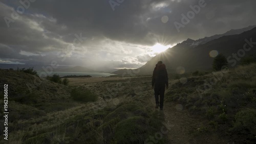 Hiker walking toward a sunburst over a mountain lake at sunset in nature on a stormy day