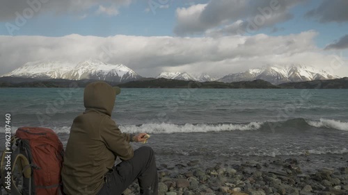 Hiker sitting by a mountain lake with waves looking at snow-capped peaks in nature