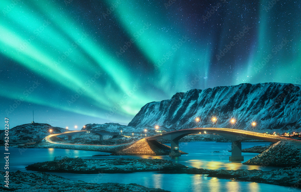 Fototapeta premium Bridge and northern lights over snowy mountains. Lofoten islands, Norway. Aurora borealis and reflection in water. Winter landscape with starry sky, polar lights, rocks, road, sea, city illumination