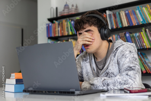 Foto Stressed and exhausted teenage student rubbing tired eyes while studying with laptop and books, preparation for difficult exams, academic pressure, ADHD concept