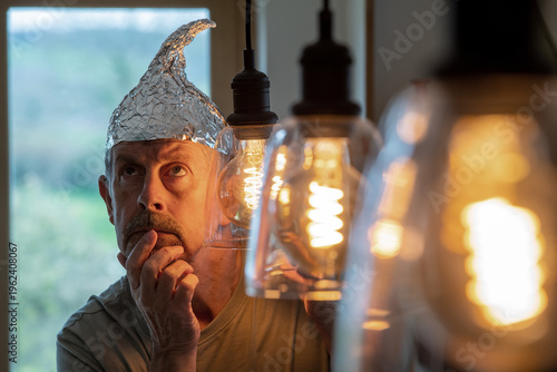 Tinfoil hat picture. A man wearing a tinfoil hat looks at his laptop with a worried look on his face. Paranoia and conspiracy theory. Alien abduction and government cover up. Mind control and brainwas