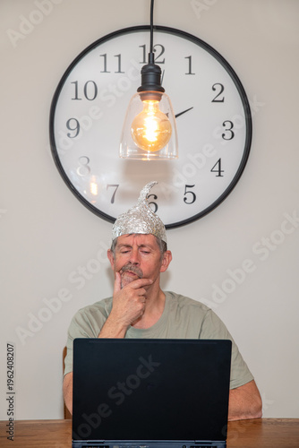 Tinfoil hat picture. A man wearing a tinfoil hat looks skeptically at his laptop. Paranoia and conspiracy theory. Alien abduction and government cover up. Mind control and brainwashing.