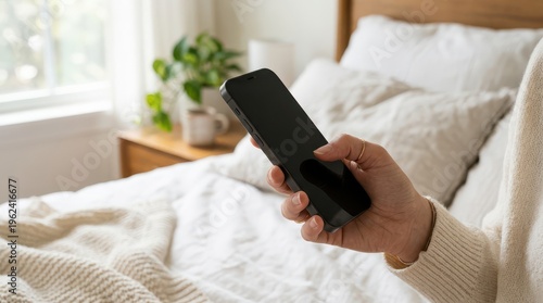 Hand holding smartphone with black screen in front of bed