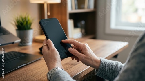 Person using smartphone at desk near laptop and window