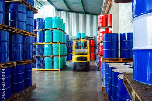 Industrial warehouse storage with rows of stacked blue, teal, and red oil barrels and a blurred yellow forklift moving through the aisle. Chemical logistics and fuel distribution concept.
