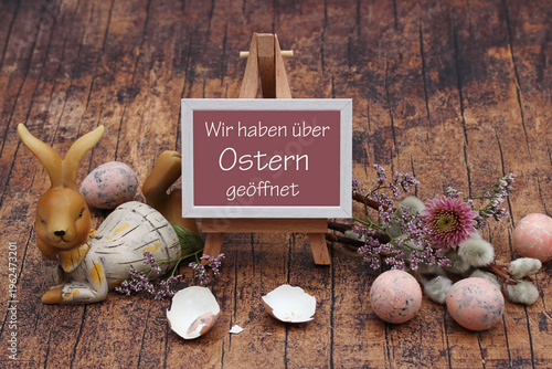 Liegende Hasenfigur neben kleiner Staffelei-Schild mit dem Text 'Wir haben über Ostern geöffnet', umgeben von zerbrochenen Eierschalen, Osterzweigen und dekorativen Eiern auf Holzboden.