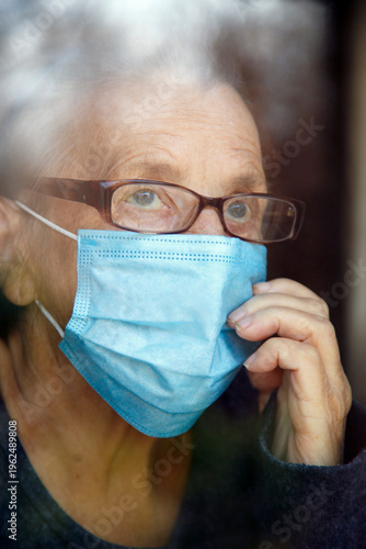 Senior woman wearing face mask looking out of glass window during covid - 1 9 pandemic. Portrait of an elderly woman in quarantine wearing a mask.