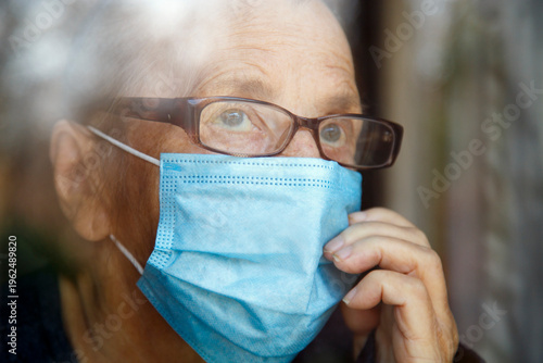 Senior woman wearing face mask looking out of glass window during covid - 1 9 pandemic. Portrait of an elderly woman in quarantine wearing a mask.