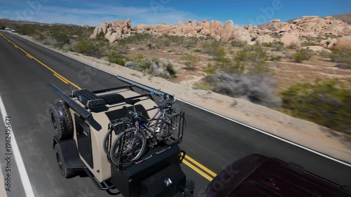 A teardrop camper trailer is towed by an SUV along a road in Joshua Tree National Park with desert views in California under a clear sky.
