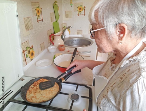 An elderly woman prepares pancakes on a gas stove in the kitchen.
