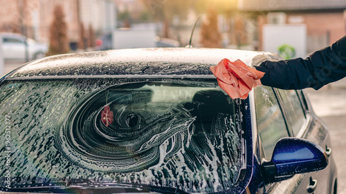 Hand cleaning car windshield with sponge and soap foam, washing vehicle outdoors, car care, maintenance and cleaning concept in warm sunlight