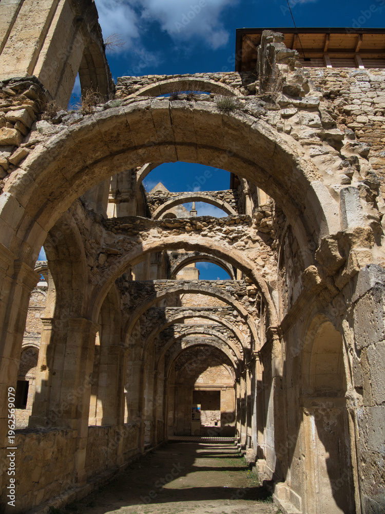 Fototapeta premium Long perspective view of ancient stone archways and cloisters - Architectural ruins of a historic monastery or abbey with sunlight streaming through the open arches and blue sky above.