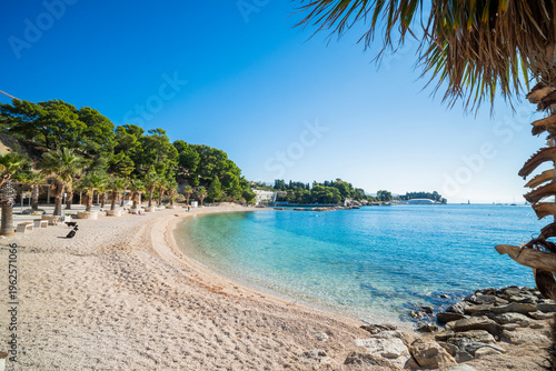 Scenic view of Kasjuni Beach in Split, Croatia. Turquoise Adriatic sea, pebble shore with palm trees and green pine forest under a clear blue sky.