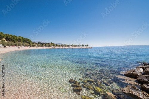 Wide-angle view of Znjan beach in Split, Croatia. Crystal clear turquoise Adriatic water with a pebble shore and green trees under a bright blue sky.