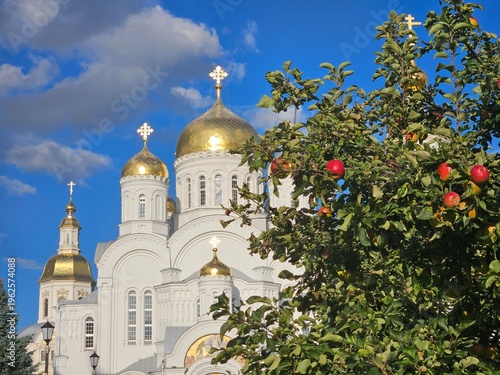 Church with a gold dome and two crosses on top. The sky is blue and there are clouds. A tree with apples on it is in front of the church