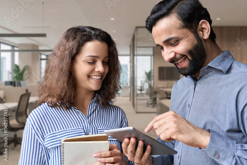 Diverse friendly coworkers colleagues indian male manager and latin female employee talking in office using tablet device, having fun and laughing together discussing project.