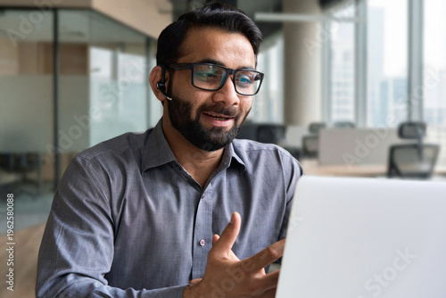 Indian business man speaking having virtual meeting on laptop. Professional remote online customer service support manager wearing headset talking consulting on video call at home office call center.