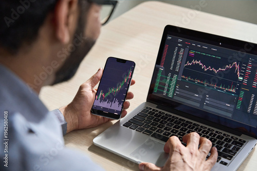 Business man trader investor analyst using mobile phone app analytics for cryptocurrency financial market analysis, trading data index chart graph on smartphone and laptop screen. Over shoulder view