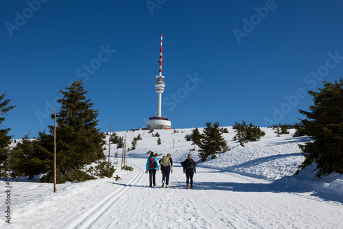Praded, Jeseniky mountains, Czech Republic, Czechia. Hikers are walking in the nature towards television and communications tower and transmitter. Hiking during sunny winter. White snow and blue sky.
