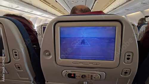 Man is sitting in an airplane with a screen in front of him. The screen is showing a video of a runway in airplane