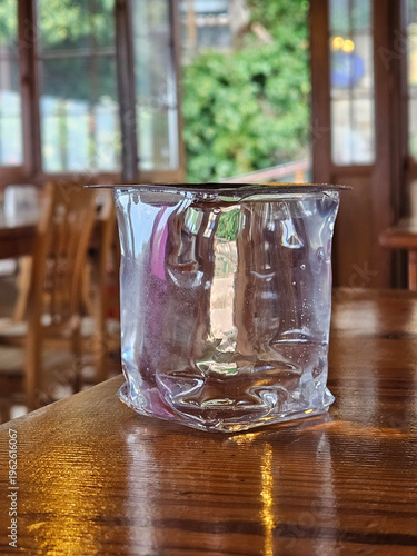 Wallpaper Mural Clear plastic cup with a plastic lid sits on a wooden table. The cup is half full and has a plastic lid on top. The table is surrounded by chairs, and there is a potted plant in the background Torontodigital.ca
