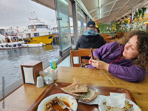 Wallpaper Mural Woman is sitting at a table with a plate of food in front of her. She is looking at her phone. The table is near a window that overlooks a body of water Torontodigital.ca