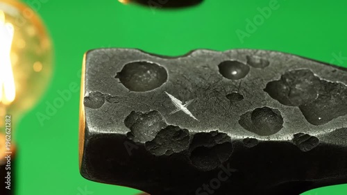 Closeup macro shot of a blacksmith hammer striking metal, showing sparks and textured surface with a vintage light bulb in background, industrial process