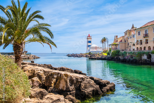 Santa Marta lighthouse and rocky coastline with palm tree in Cascais, Portugal. White and blue lighthouse, clear sea water and historic buildings in bright sunlight
