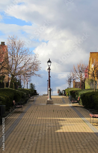Empty pedestrian walkway with benches and lamppost under dramatic sky.