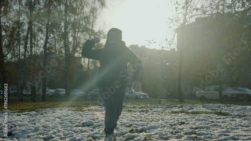 Backlit hooded figure walking across snow. Tense late afternoon light with lens flare, bare trees and parked cars in background, footprints on frozen grass, slow stride suggesting snatcher or prowler,