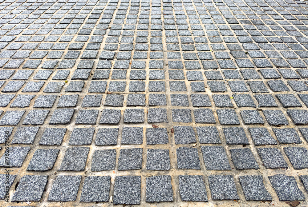 Fototapeta premium Gray stone square tiles arranged in a grid pattern fill the ground surface with sand and small debris between the stones creating a textured and geometric appearance