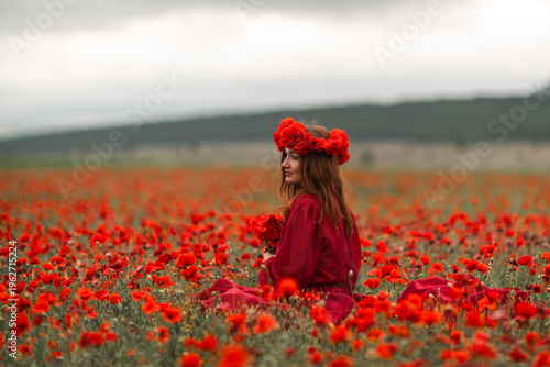 Woman poppy field sitting amidst vibrant red flowers wearing a crown in misty landscape.