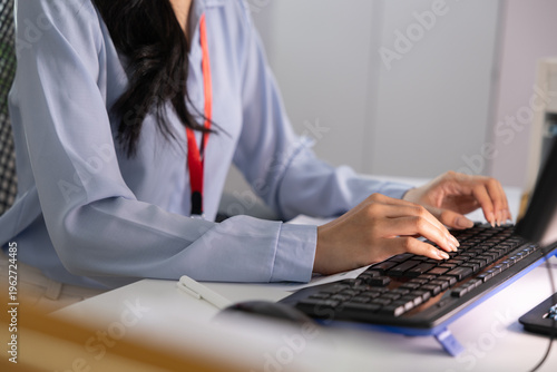 Focused professional woman working at modern office desk typing on computer keyboard showing diligent concentration while wearing light blue shirt and red lanyard