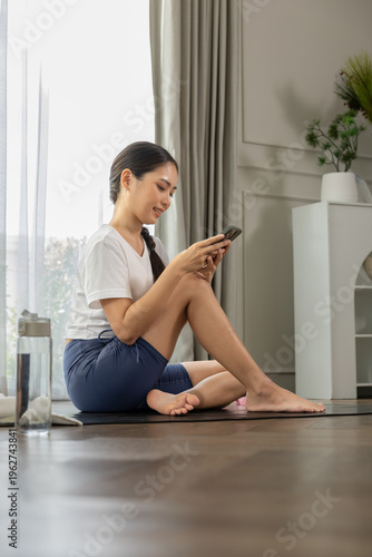 Happy asian woman sitting on yoga mat near vertical window holding smartphone feeling relaxed wearing white shirt and blue shorts resting after workout at home enjoying peaceful morning routine Free Download Happy asian woman sitting on yoga mat near vertical window holding smartphone feeling relaxed wearing white shirt and blue shorts resting after workout at home enjoying peaceful morning routine - GFXLoad