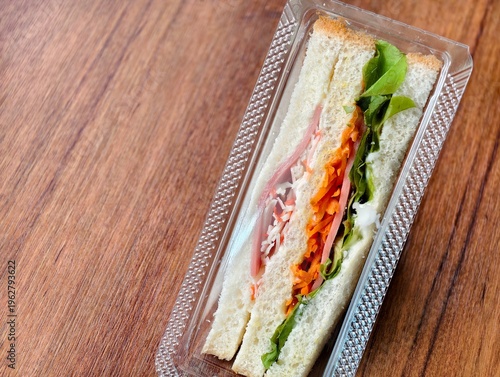 Top view of fresh club sandwich packed with lettuce, carrot, ham, and crab stick in a clear takeaway plastic box on a wooden table. Quick lunch or snack concept