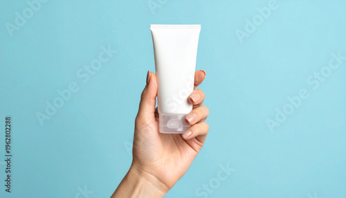 Close-up of a woman's hand holding a white blank cosmetic tube mockup against a solid light blue background. Minimalist skincare packaging concept
