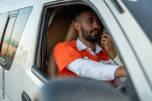Driver worker male adult safety vest radio vehicle window steering wheel uniform focused worker speaks into two way radio inside vehicle, holding steering wheel, calm and alert
