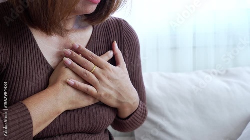 Middle-aged woman clutching her chest in pain, suffering from a sudden heart attack or chest tightness while sitting on a sofa at home. Concept of cardiovascular disease and emergency healthcare