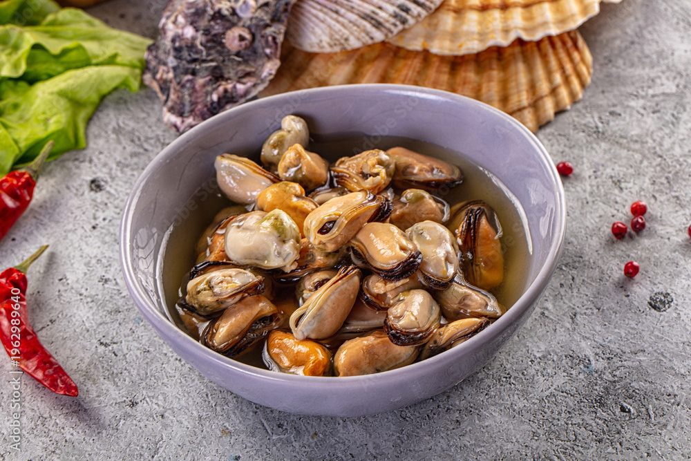 Fototapeta premium Marinated mussels in a bowl with chili peppers, lettuce, and seashells on a rustic grey background.