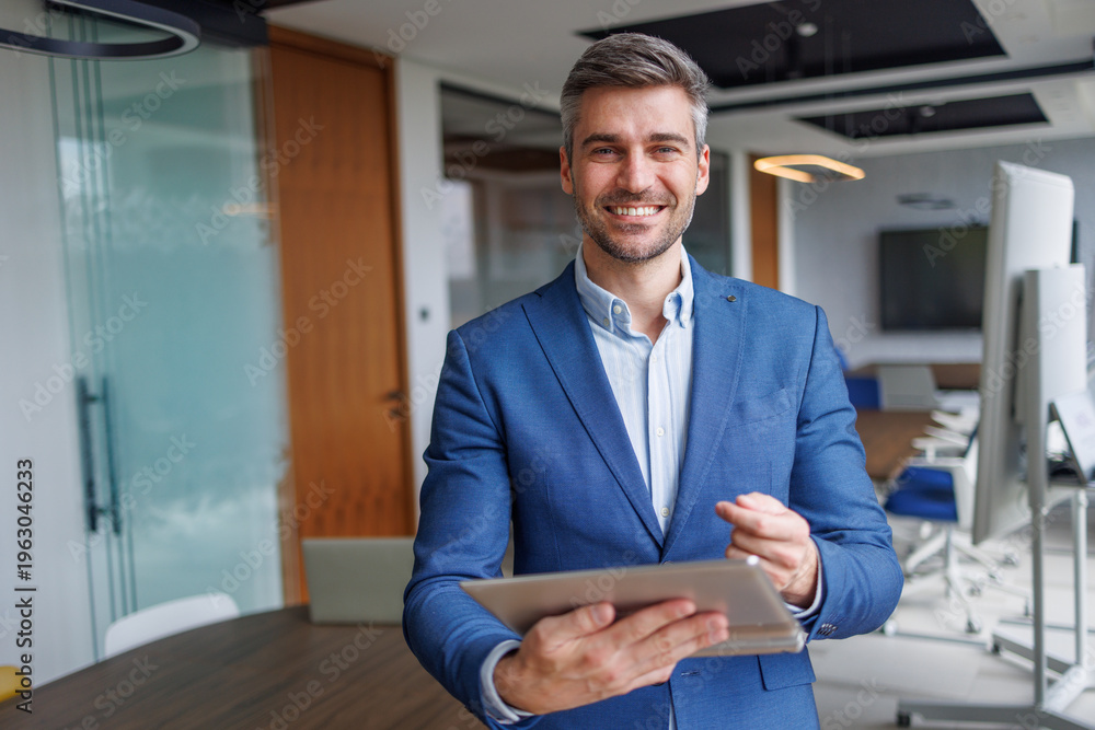 Fototapeta premium Confident businessman smiling, holding a tablet while standing in a modern office, representing success and technology