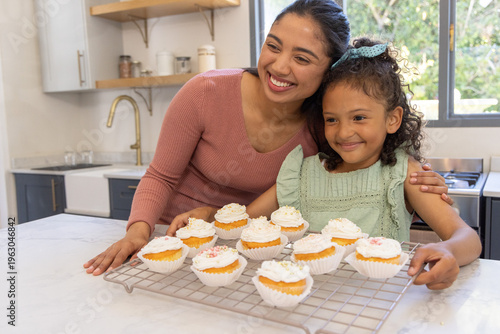 Mother and child smiling and presenting frosted cupcakes on cooling rack on marble countertop