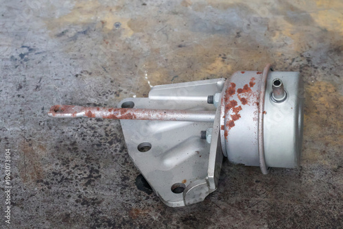 Photos A metal turbocharger wastegate actuator with a rusty control rod and mounting bracket is shown from a top-down view on a dirty industrial floor for engine part replacement
